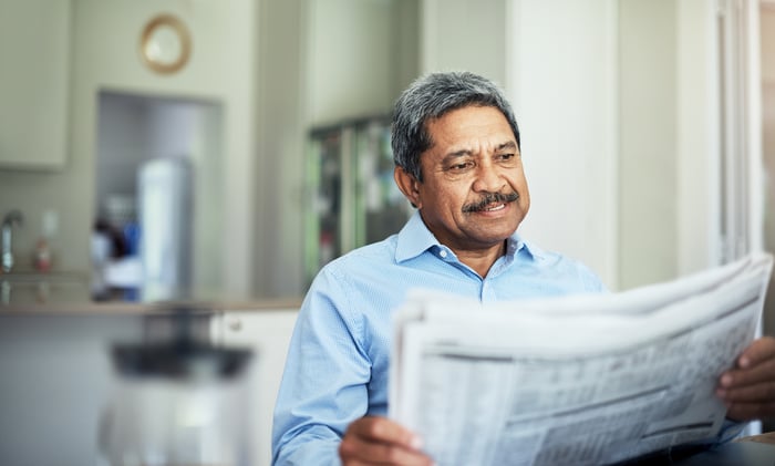Smiling person reading financial newspaper in a kitchen.