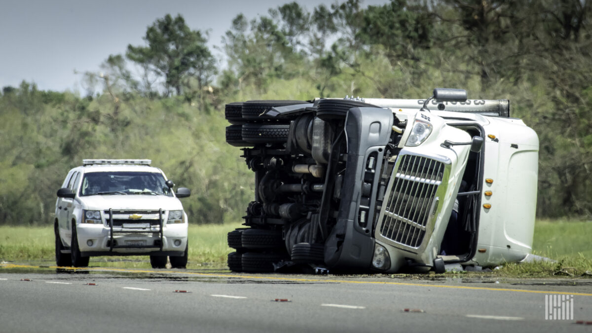 A white commercial truck sits on its side after a crash with a police car parked behind it.