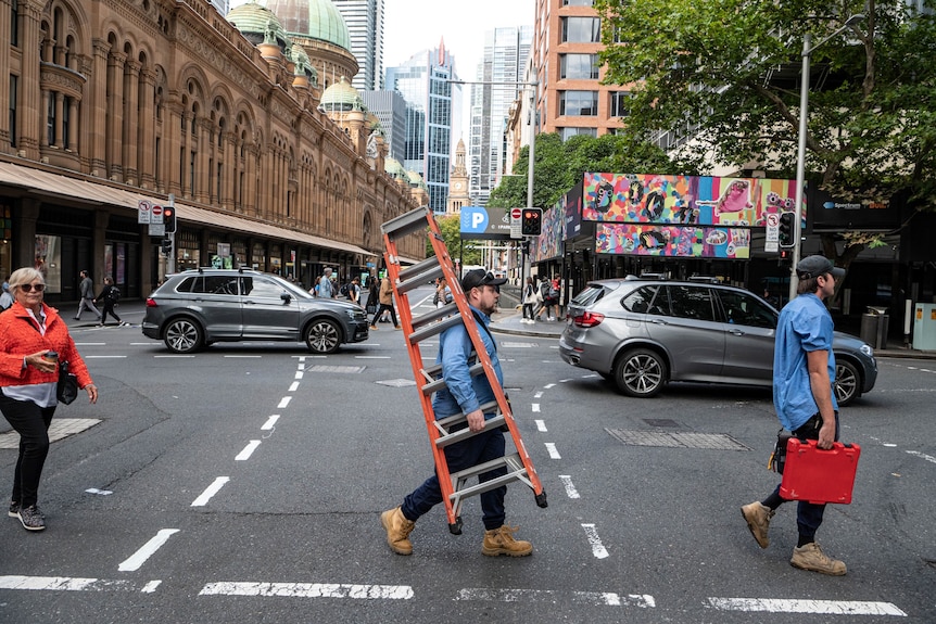 Three people cross a road in a CBD. Two are tradesmen, with one carrying an orange ladder. A woman is walking behind them.