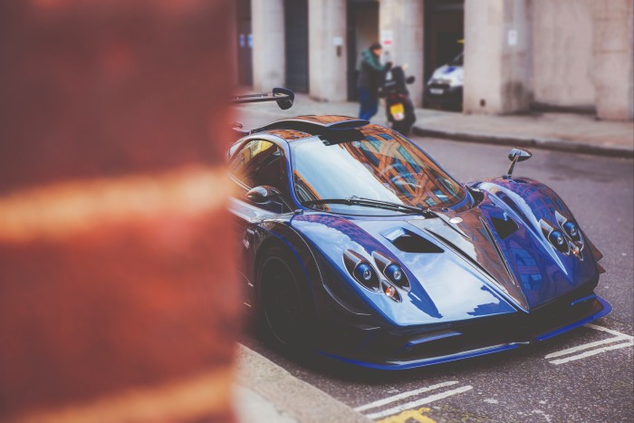 A high-performance blue Pagani Zonda is seen parked along an urban street, with its reflective body showcasing the architecture around it. The focus on the car highlights its elegant curves and sporty features, while a person on a motorbike and a building entrance can be seen in the background